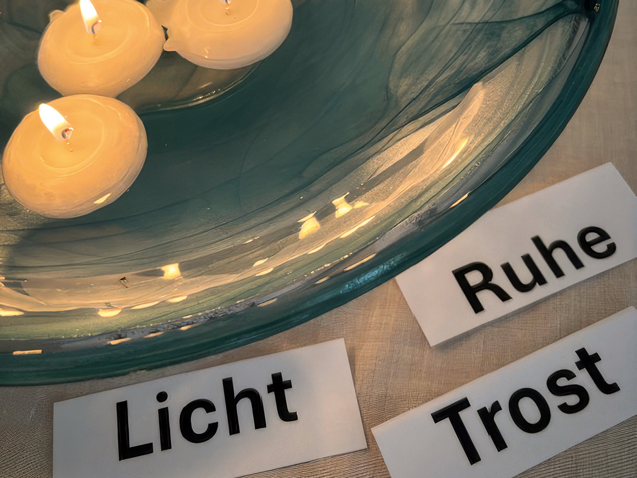 Eine flache Wasserschale, in der brennende Schwimmkerzen schwimmen, davor auf einem Tisch liegen Kärtchen mit Worten wie Licht, Ruhe und Trost